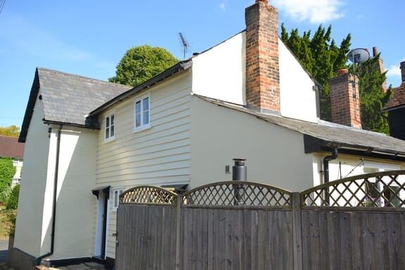 Cream-painted cottage with weatherboarded and rendered walls, slate roofs and red brick chimneys behind a fence