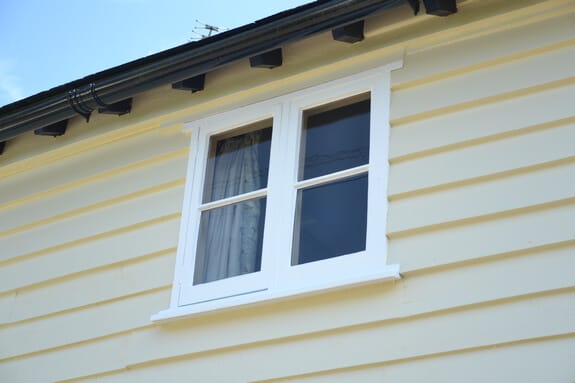 White-painted two-light timber casement window in cream weatherboarding beneath black-painted rafter ends