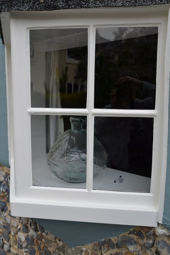 Small four-pane timber window painted white above flint walling, a glass vase on the sill inside