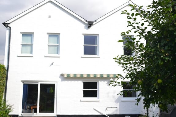 White-painted rear elevation with twin gables, sash windows and a striped awning, a fruit tree in the foreground