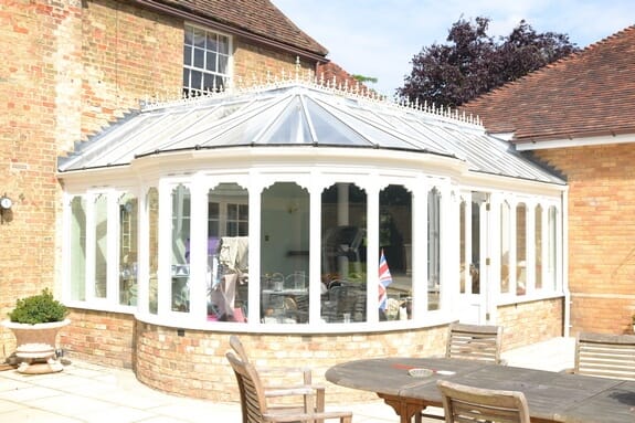 White conservatory with curved glazed sides and a glass roof with ornamental cresting, on a brick base
