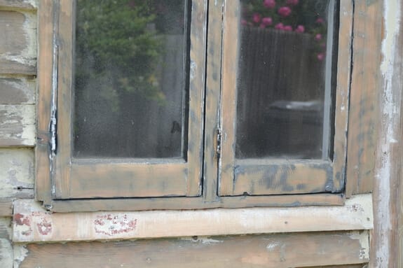 Close-up of a two-light timber casement window with paint worn back to bare wood above a weathered sill board