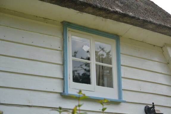 Small casement window with a blue-painted frame in white weatherboarding beneath thatched eaves