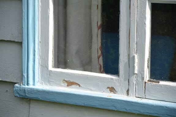 Close-up of flaking white paint on a casement window's bottom rail, next to its blue-painted frame