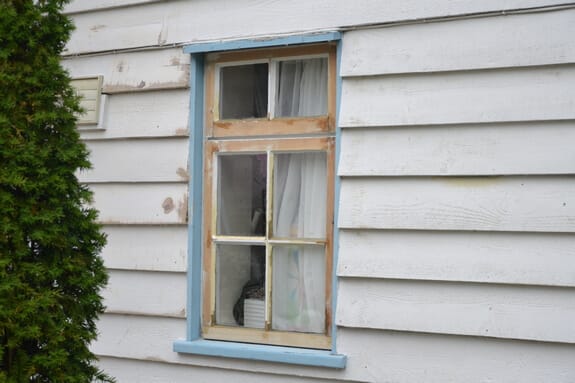 Casement window stripped back to bare timber inside its blue-painted frame in white weatherboarding