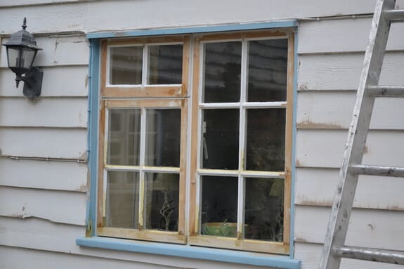 Pair of casement windows stripped to bare wood in white weatherboarding, with a wall lantern and ladder