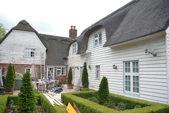 Thatched, weatherboarded cottage and garden with box hedges and topiary, ladders up against the walls
