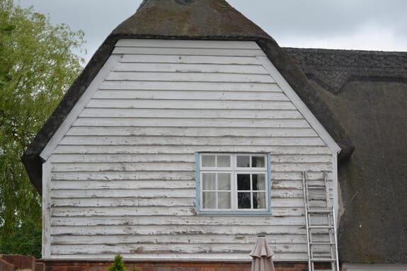 Thatched cottage gable with flaking white paint on the weatherboard cladding, a casement window and a ladder