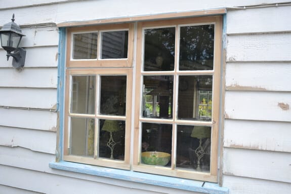 Worn timber casement window with a pale blue surround in white weatherboard cladding beside a wall lantern