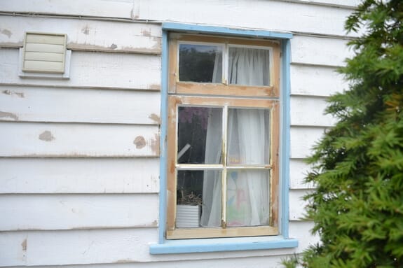 Weathered timber casement window with a pale blue surround in weatherboard cladding next to a louvred vent