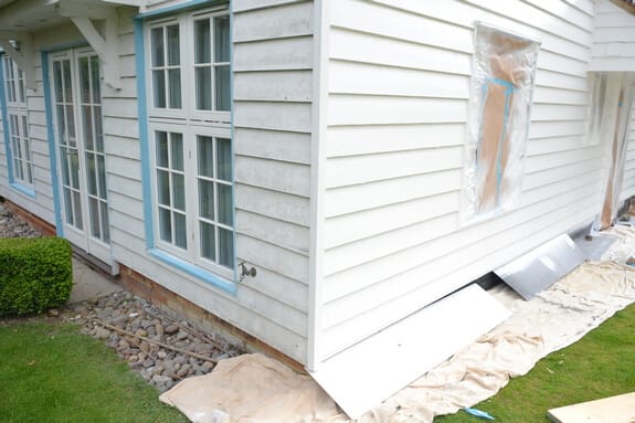 White weatherboard wall with pale blue-framed windows, one masked in plastic, and dust sheets along the base