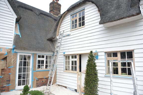 Thatched cottage with ladders up the walls and brown paper and blue tape masking the lower windows and brickwork