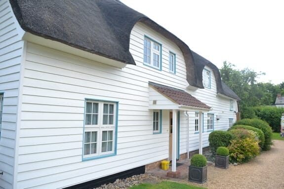 Front of the thatched cottage with white weatherboarding, pale blue window frames and a tiled porch