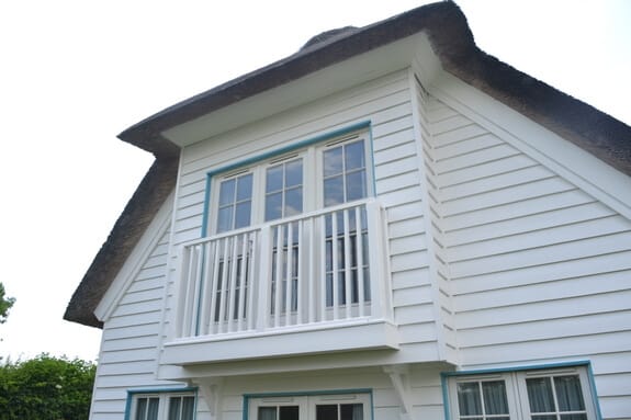 Weatherboarded gable with a first-floor balcony, white balustrade and pale blue window frames under thatch