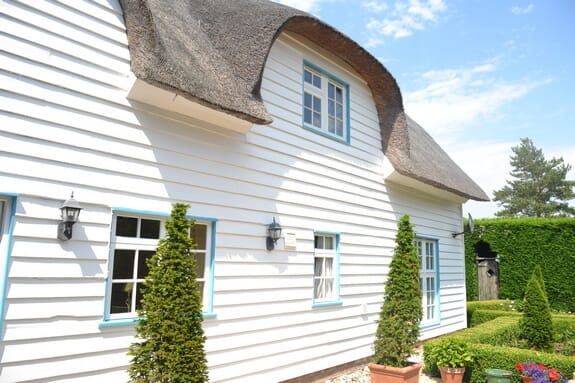 Side of the thatched cottage with white weatherboarding, pale blue windows, wall lanterns and potted conifers