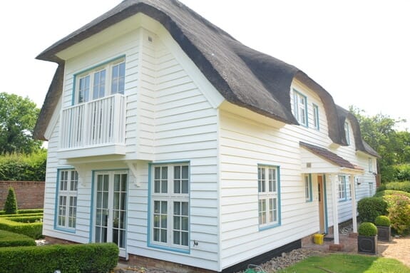 Thatched cottage with white weatherboarding, a balcony gable, French doors and pale blue windows