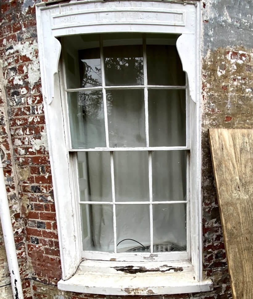 A six-over-six sash window under a curved hood, its paint and sill weathered
