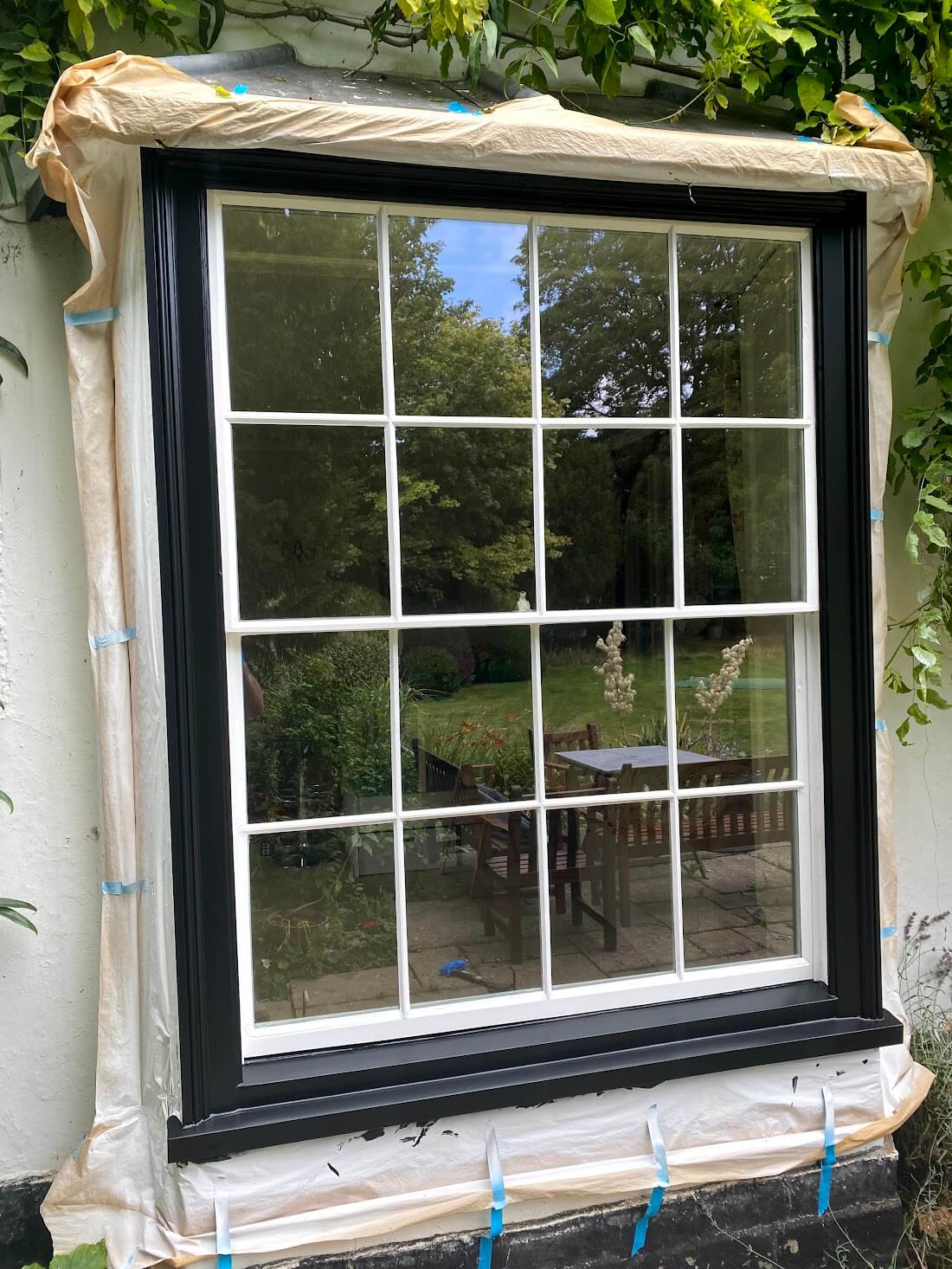 Six-over-six sash window with white sashes and a black-painted frame, masked off with paper and blue tape for painting