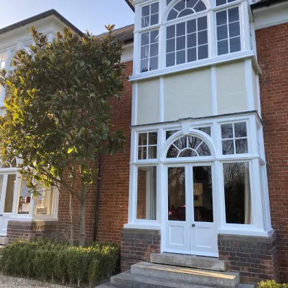 A red brick house with large white-framed sash windows, recently restored by a sash windows repair Bury St Edmunds specialist, stands beside a leafy tree and low green hedges.