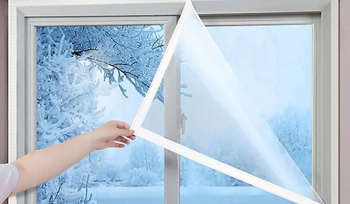 Hand fitting clear insulating film over a white window frame, with snow-covered trees outside